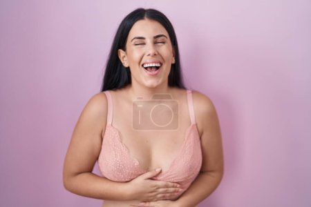 Foto de Mujer hispana joven con sujetador rosa sonriendo y riendo en voz alta porque divertida broma loca con las manos en el cuerpo. - Imagen libre de derechos