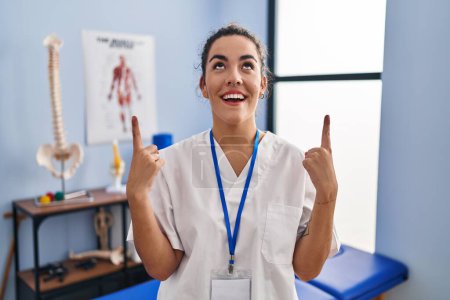Téléchargez les photos : Young hispanic woman working at rehabilitation clinic amazed and surprised looking up and pointing with fingers and raised arms. - en image libre de droit
