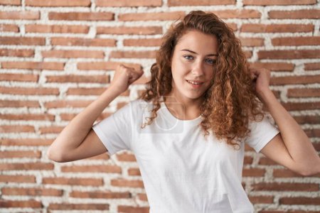 Téléchargez les photos : Jeune femme caucasienne debout sur un fond de mur de briques montrant les muscles des bras souriant fier. concept de fitness. - en image libre de droit