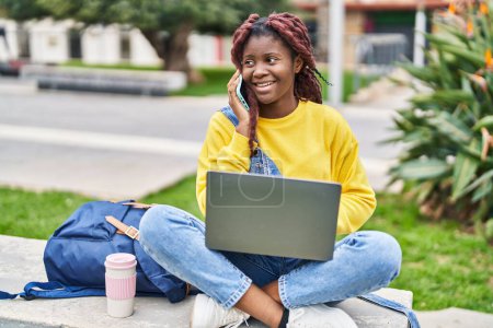 Téléchargez les photos : Étudiante afro-américaine utilisant un ordinateur portable parlant sur smartphone au parc du campus - en image libre de droit
