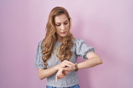 Foto de Hermosa mujer rubia de pie sobre fondo rosa comprobar la hora en el reloj de pulsera, relajado y seguro - Imagen libre de derechos