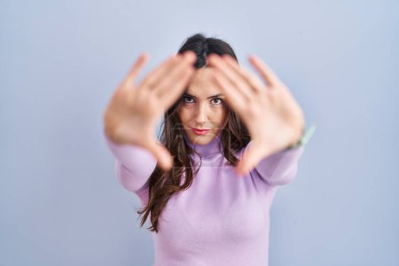 Téléchargez les photos : Jeune femme brune debout sur fond bleu faisant cadre en utilisant les mains paumes et les doigts, perspective de la caméra - en image libre de droit