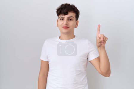 Téléchargez les photos : Jeune homme non binaire portant t-shirt blanc décontracté montrant et pointant vers le haut avec le doigt numéro un tout en souriant confiant et heureux. - en image libre de droit