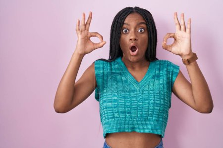 Foto de Joven afroamericano con trenzas de pie sobre fondo rosa mirando sorprendido y sorprendido haciendo símbolo de aprobación ok con los dedos. expresión loca - Imagen libre de derechos