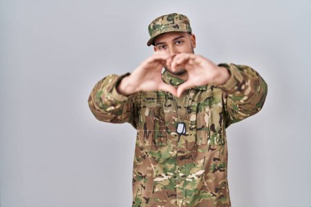 Foto de Young hispanic man wearing camouflage army uniform smiling in love doing heart symbol shape with hands. romantic concept. - Imagen libre de derechos
