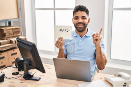 Foto de Hombre hispano con barba trabajando en la oficina con laptop sosteniendo banner de agradecimiento sonriendo feliz señalando con la mano y el dedo a un lado - Imagen libre de derechos