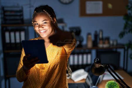 Téléchargez les photos : Femme afro-américaine avec des tresses travaillant au bureau la nuit avec tablette approuvant faire un geste positif avec la main, pouces levés souriant et heureux pour le succès. geste gagnant. - en image libre de droit