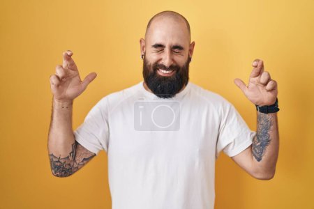 Téléchargez les photos : Jeune homme hispanique avec barbe et tatouages debout sur fond jaune geste doigt croisé souriant avec espoir et les yeux fermés. chance et concept superstitieux. - en image libre de droit