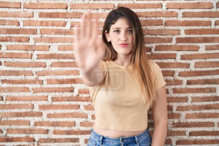 Photo for Young brunette woman standing over bricks wall doing stop sing with palm of the hand. warning expression with negative and serious gesture on the face. - Royalty Free Image
