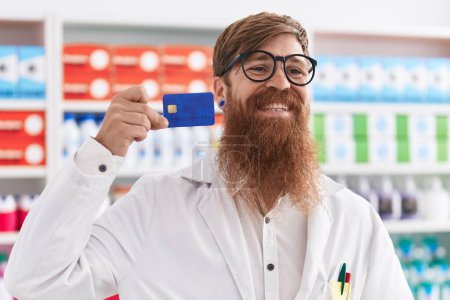 Foto de Joven pelirroja hombre farmacéutico sonriendo confiado celebración de la tarjeta de crédito en la farmacia - Imagen libre de derechos
