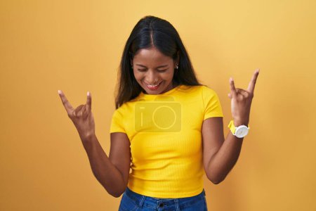 Foto de Mujer india joven de pie sobre fondo amarillo gritando con expresión loca haciendo símbolo de roca con las manos arriba. Estrella de la música. concepto de música pesada. - Imagen libre de derechos