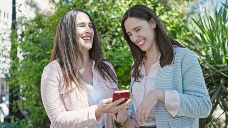 Foto de Dos mujeres sonriendo confiadas usando smartphone en el parque - Imagen libre de derechos