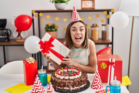 Foto de Mujer hispana celebrando cumpleaños con pastel sosteniendo regalo sonriendo y riendo en voz alta porque divertida broma loca. - Imagen libre de derechos