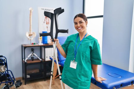 Photo for Young beautiful hispanic woman pysiotherapist smiling confident holding crutches at rehab clinic - Royalty Free Image