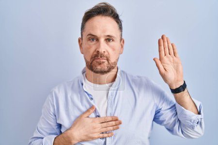 Photo for Middle age caucasian man standing over blue background swearing with hand on chest and open palm, making a loyalty promise oath - Royalty Free Image