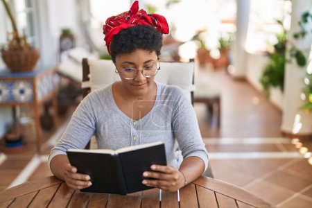 Téléchargez les photos : Femme afro-américaine livre de lecture assis sur la table à la maison terrasse - en image libre de droit