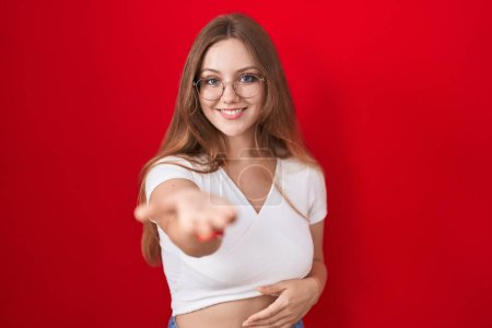 Téléchargez les photos : Jeune femme caucasienne debout sur fond rouge souriant joyeuse offrant main de paume donnant assistance et acceptation. - en image libre de droit