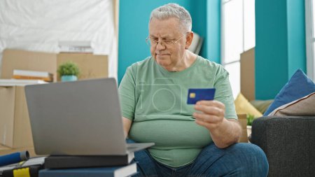 Téléchargez les photos : Homme aux cheveux gris du Moyen Âge faisant du shopping avec un ordinateur portable et une carte de crédit assis sur un canapé dans une nouvelle maison - en image libre de droit