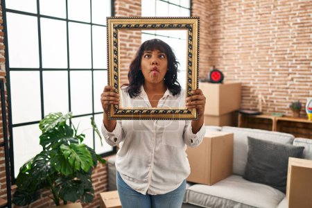Photo for Hispanic woman at new home holding empty frame making fish face with mouth and squinting eyes, crazy and comical. - Royalty Free Image
