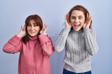 Téléchargez les photos : Mère et fille debout sur fond bleu essayant d'entendre les deux mains sur le geste de l'oreille, curieux de commérages. problème auditif, sourd - en image libre de droit