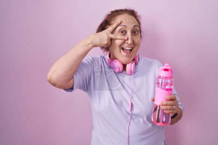 Téléchargez les photos : Femme âgée portant des vêtements de sport et des écouteurs faisant symbole de paix avec les doigts sur le visage, souriant joyeuse montrant la victoire - en image libre de droit