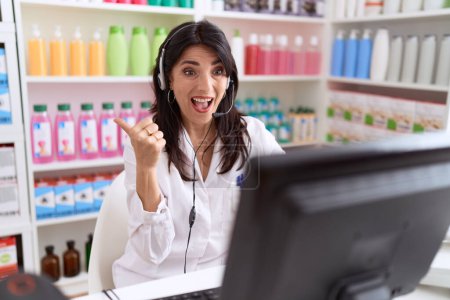 Téléchargez les photos : Moyen Age femme hispanique travaillant à la pharmacie pharmacie en utilisant casque et ordinateur pointant pouce vers le côté souriant heureux avec bouche ouverte - en image libre de droit