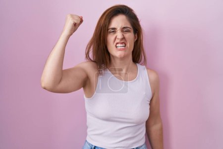 Téléchargez les photos : Femme brune debout sur fond rose en colère et fou levant le poing frustré et furieux tout en criant de colère. rage et concept agressif. - en image libre de droit