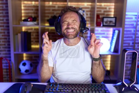 Téléchargez les photos : Middle age man with beard playing video games wearing headphones gesturing finger crossed smiling with hope and eyes closed. luck and superstitious concept. - en image libre de droit