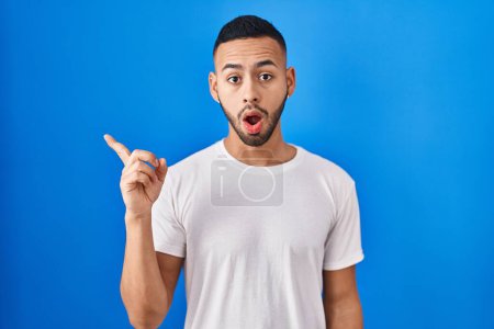 Téléchargez les photos : Young hispanic man standing over blue background surprised pointing with finger to the side, open mouth amazed expression. - en image libre de droit