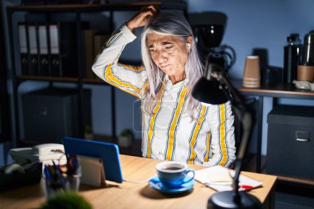 Téléchargez les photos : Femme d'âge moyen avec les cheveux gris travaillant au bureau la nuit confondre et s'interroger sur la question. incertain avec doute, pensant la main sur la tête. concept coûteux. - en image libre de droit