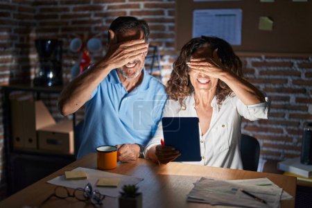 Photo for Middle age hispanic couple using touchpad sitting on the table at night smiling and laughing with hand on face covering eyes for surprise. blind concept. - Royalty Free Image