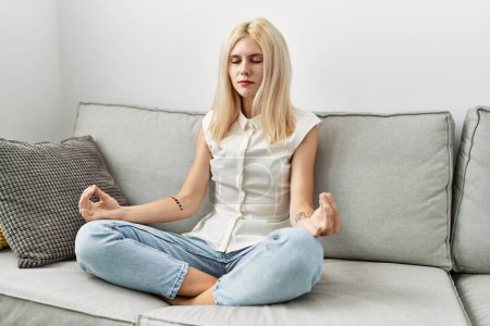 Foto de Mujer rubia joven haciendo ejercicio de yoga sentado en el sofá en casa - Imagen libre de derechos