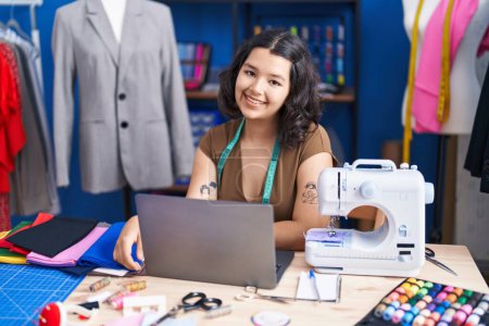 Téléchargez les photos : Jeune femme tailleur souriant confiant en utilisant un ordinateur portable au studio de couture - en image libre de droit