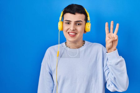 Téléchargez les photos : Personne non binaire écoutant de la musique à l'aide d'écouteurs montrant et pointant vers le haut avec les doigts numéro trois tout en souriant confiant et heureux. - en image libre de droit