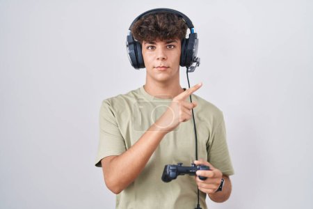 Téléchargez les photos : Adolescent hispanique jouant contrôleur de tenue de jeu vidéo pointant avec le doigt de la main sur le côté montrant la publicité, visage sérieux et calme - en image libre de droit