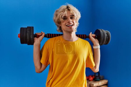 Foto de Joven hombre rubio sonriendo confiado usando el entrenamiento con pesas en el centro deportivo - Imagen libre de derechos