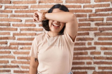 Photo for Young hispanic woman standing over bricks wall covering eyes with arm smiling cheerful and funny. blind concept. - Royalty Free Image