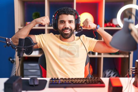 Photo for Hispanic man with beard playing video games with headphones showing arms muscles smiling proud. fitness concept. - Royalty Free Image