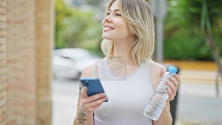 Téléchargez les photos : Jeune femme blonde utilisant un smartphone tenant une bouteille d'eau dans la rue - en image libre de droit