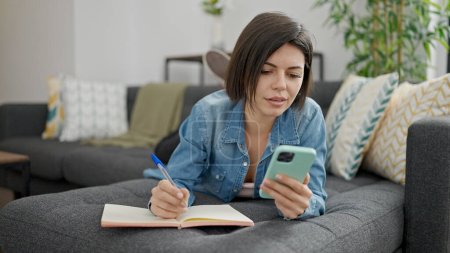 Téléchargez les photos : Jeune femme caucasienne utilisant l'écriture smartphone sur ordinateur portable à la maison - en image libre de droit