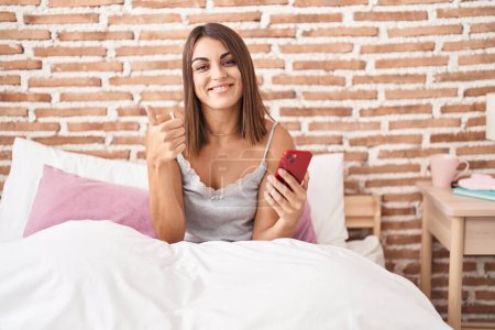 Foto de Mujer hispana joven usando teléfono inteligente sentado en la cama en casa sonriendo feliz y positivo, pulgar hacia arriba haciendo excelente y signo de aprobación - Imagen libre de derechos