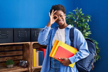 Téléchargez les photos : Femme afro-américaine portant un sac à dos étudiant et tenant des livres souriant heureux faisant signe ok avec la main sur les yeux regardant à travers les doigts - en image libre de droit