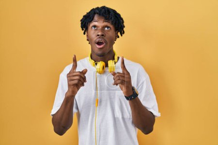 Téléchargez les photos : Jeune homme africain avec dreadlocks debout sur fond jaune étonné et surpris regardant vers le haut et pointant avec les doigts et les bras levés. - en image libre de droit
