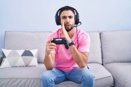 Photo for Hispanic young man playing video game holding controller sitting on the sofa hand on mouth telling secret rumor, whispering malicious talk conversation - Royalty Free Image
