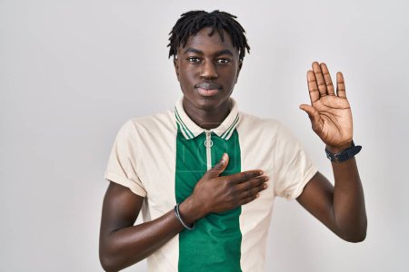 Foto de Hombre africano con rastas de pie sobre fondo aislado jurando con la mano en el pecho y la palma abierta, haciendo un juramento de lealtad promesa - Imagen libre de derechos