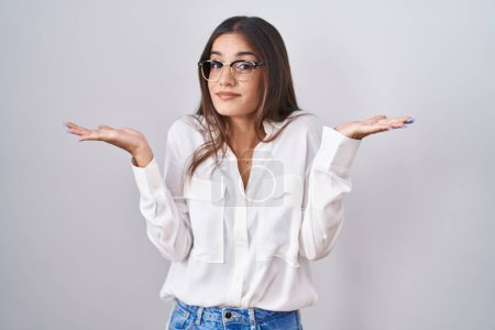 Photo for Young brunette woman wearing glasses clueless and confused expression with arms and hands raised. doubt concept. - Royalty Free Image