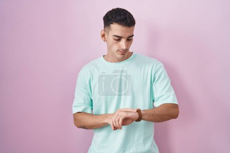 Téléchargez les photos : Bel homme hispanique debout sur fond rose vérifier l'heure sur montre-bracelet, détendu et confiant - en image libre de droit