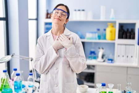 Photo for Young brunette woman working at scientist laboratory smiling with hands on chest, eyes closed with grateful gesture on face. health concept. - Royalty Free Image