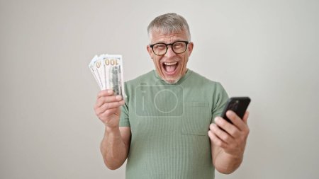 Foto de Hombre de pelo gris de mediana edad usando un teléfono inteligente que sostiene dólares sobre un fondo blanco aislado - Imagen libre de derechos