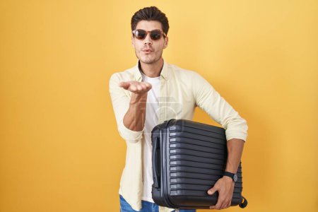 Téléchargez les photos : Jeune homme hispanique tenant une valise allant en vacances d'été en regardant la caméra souffler un baiser avec la main sur l'air étant belle et sexy. expression d'amour. - en image libre de droit